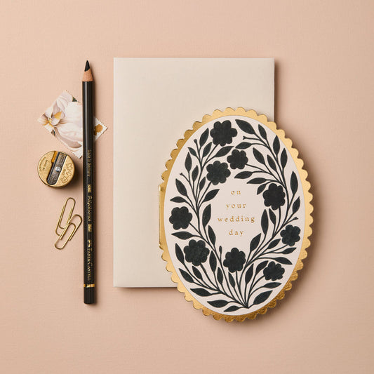 Black floral wreath illustration on oval card with gold scalloped edge, reading On Your Wedding Day