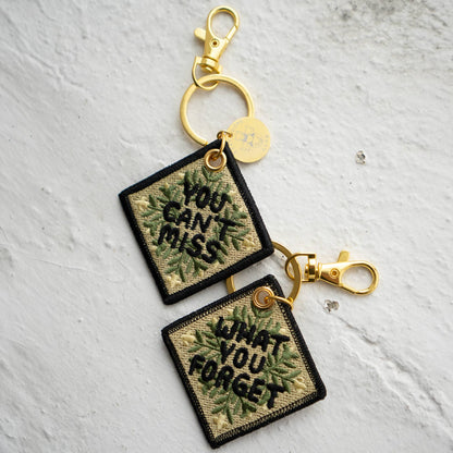 Two square embroidered keychains reading 'You Can't Miss' and 'What You Forget' in a leafy quilt-pattern design, attached with gold hardware