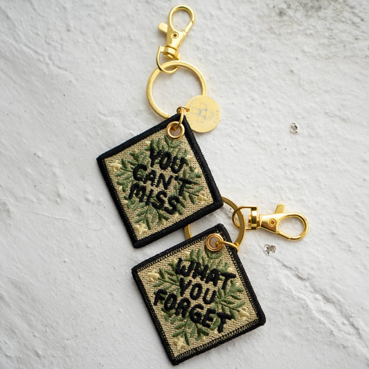 Two square embroidered keychains reading 'You Can't Miss' and 'What You Forget' in a leafy quilt-pattern design, attached with gold hardware