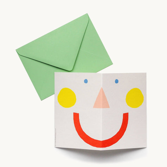 Cream greeting card folded open to reveal a large minimalist smiley face made of yellow cheeks, a peach triangle nose, blue dot eyes and a red smile, with a green envelope behind