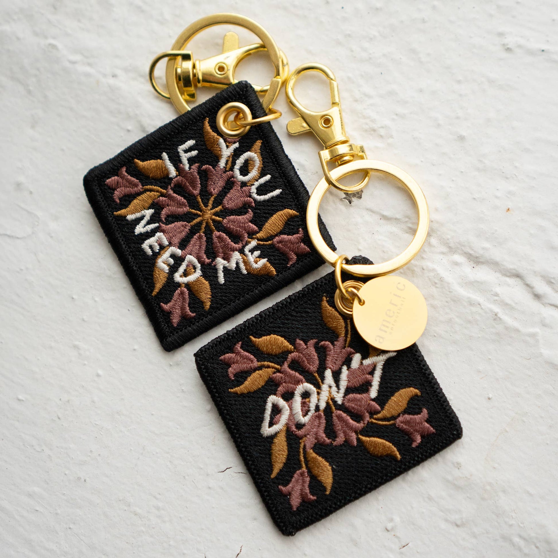 Two floral embroidered keychains reading 'If You Need Me' and 'Don't' in maroon and gold thread, attached with gold-tone clasps and a round branded charm