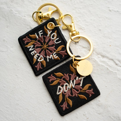 Two floral embroidered keychains reading 'If You Need Me' and 'Don't' in maroon and gold thread, attached with gold-tone clasps and a round branded charm