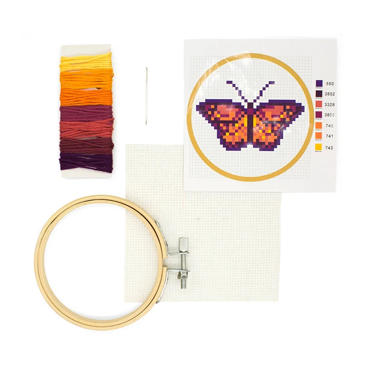 Butterfly cross stitch embroidery kit contents laid out, including colour threads, needle, canvas, bamboo hoop, and pattern card with orange and purple butterfly design.