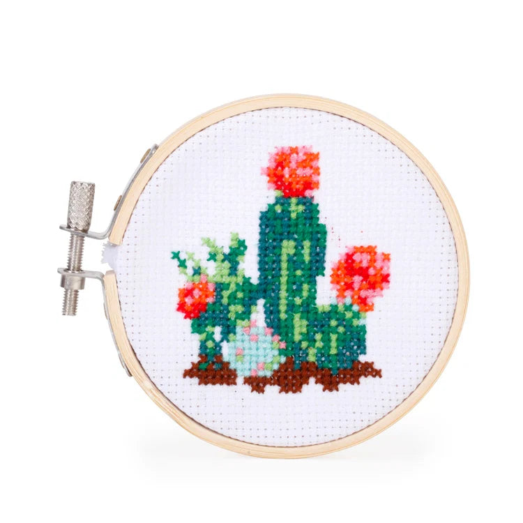 Completed cactus cross stitch pattern in a bamboo hoop, showing green cacti with pink and orange flowers on white fabric.