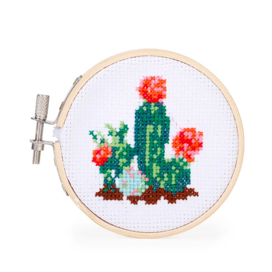 Completed cactus cross stitch pattern in a bamboo hoop, showing green cacti with pink and orange flowers on white fabric.
