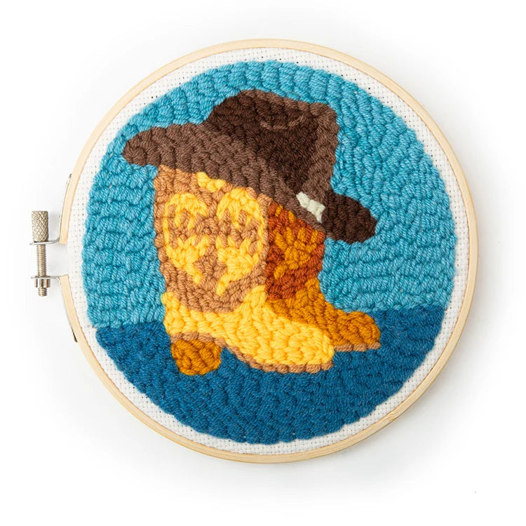 Completed Western punch needle embroidery in a bamboo hoop, showing a cowboy hat and boots design on a teal background.