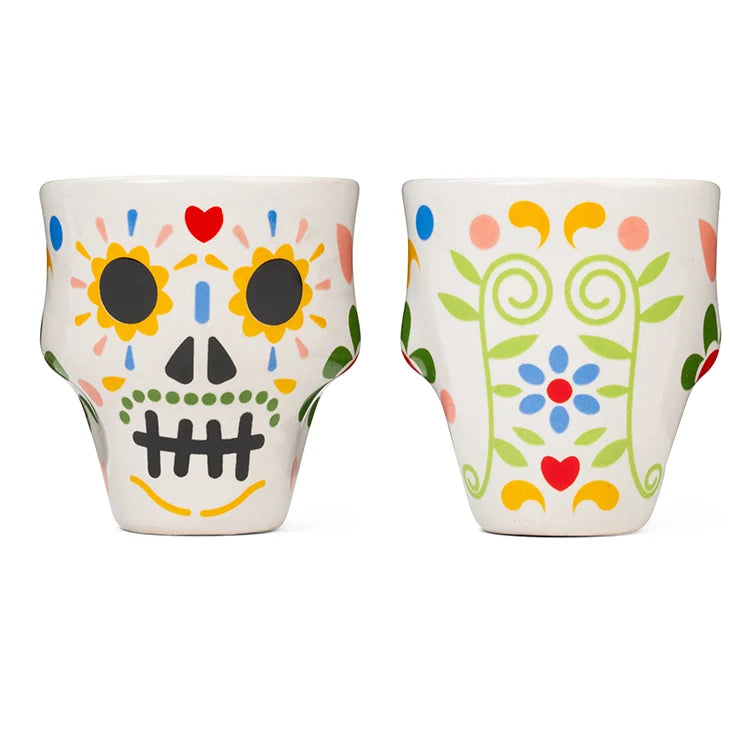Two dolomite ceramic shot glasses decorated with colourful Día de Muertos sugar skull and floral designs, standing side by side.