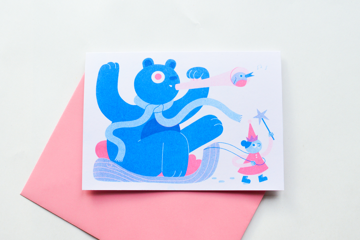 Sleigh Ride Seasonal A6 Risograph Card — illustration of a blue bear riding a sleigh pulled by a fairy, shown on a pink envelope