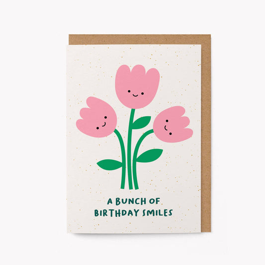 Cream greeting card with three illustrated smiling pink tulips and the text 'A Bunch of Birthday Smiles,' kraft envelope alongside