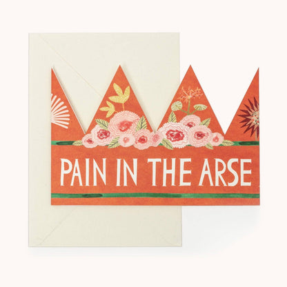 Orange party hat crown card with a pink floral band and the words 'PAIN IN THE ARSE' in white, with a cream envelope behind