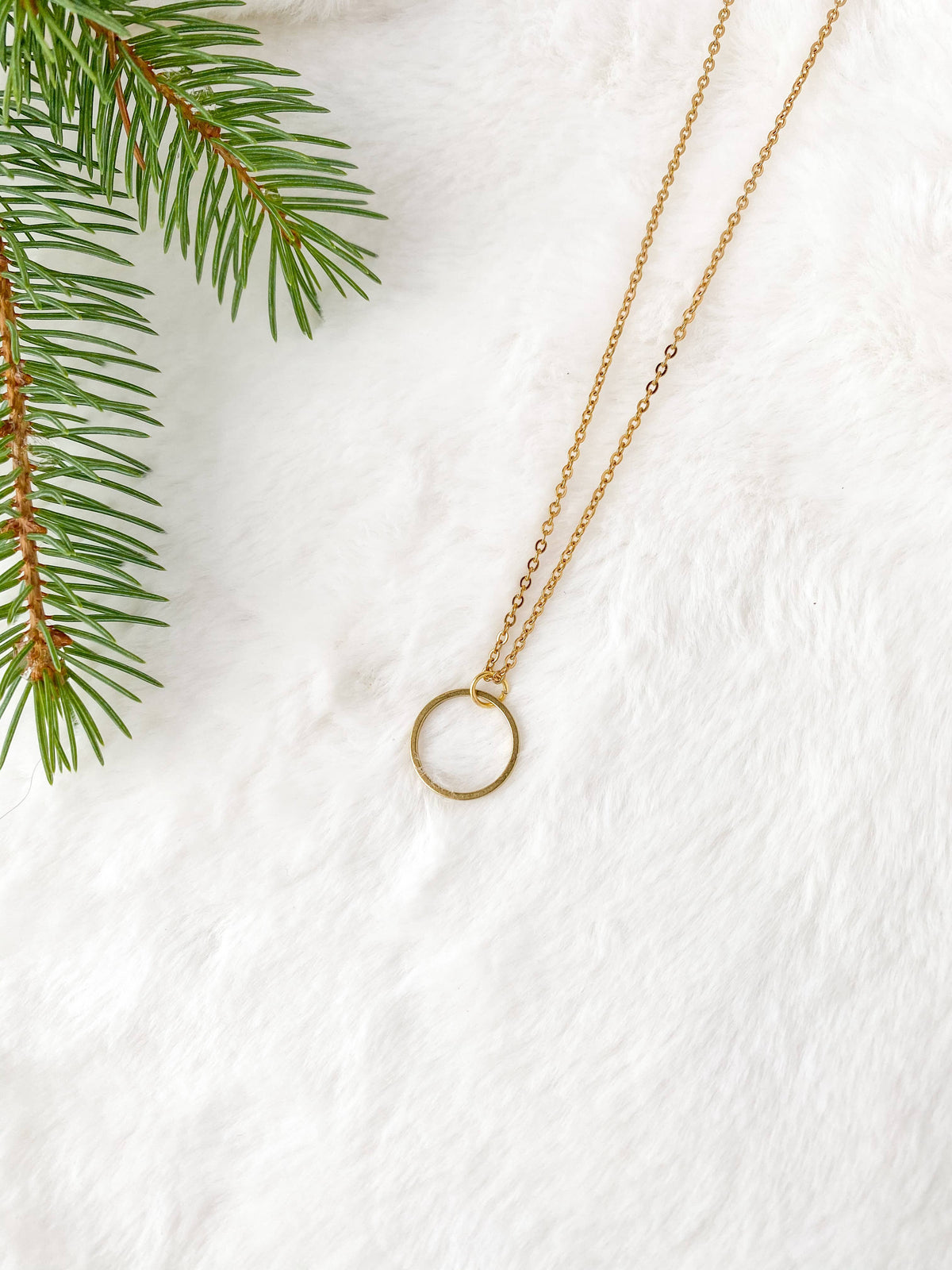 Gold-tone raw brass circle pendant necklace laid on white faux fur beside a pine branch