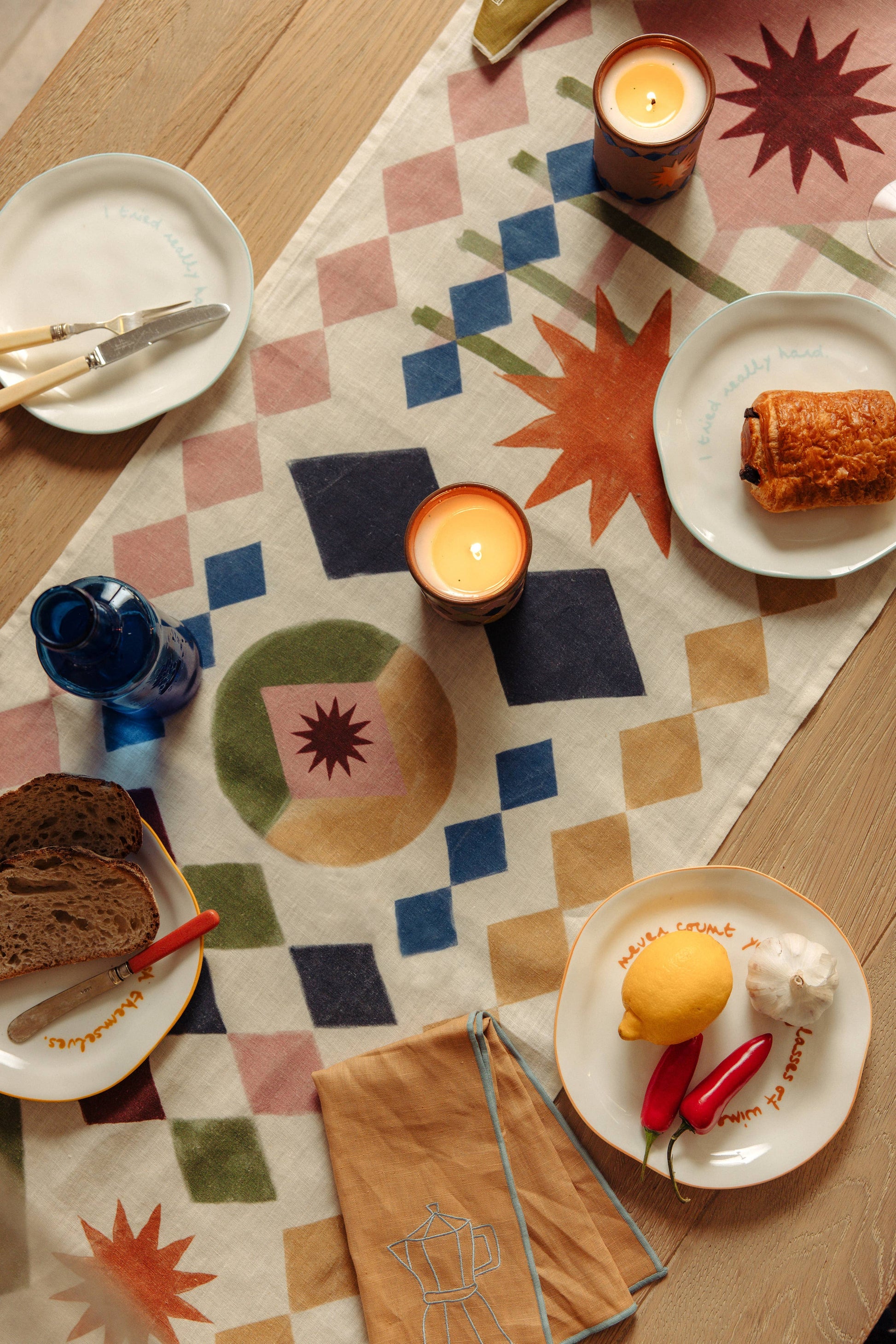 Dining table setting with plates, candles, and bread on a patterned tablecloth.