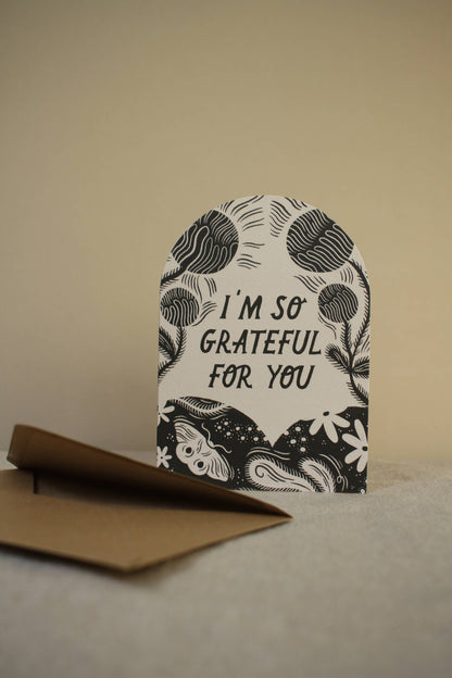 'I'm So Grateful For You' arch-shaped thank you card with black botanical wreath illustration, standing upright against a warm beige backdrop with a kraft envelope