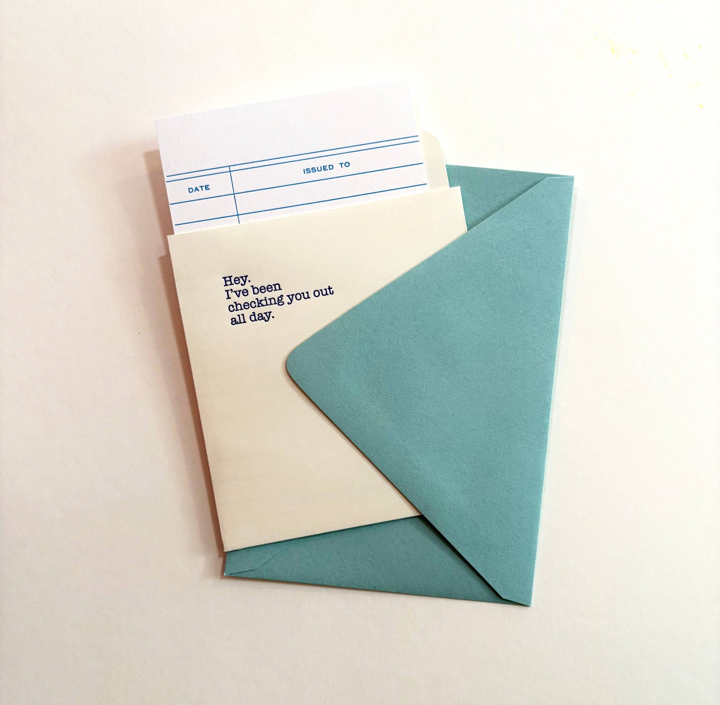Light ivory library checkout card reading Hey, I've been checking you out all day, peeking out of a teal envelope