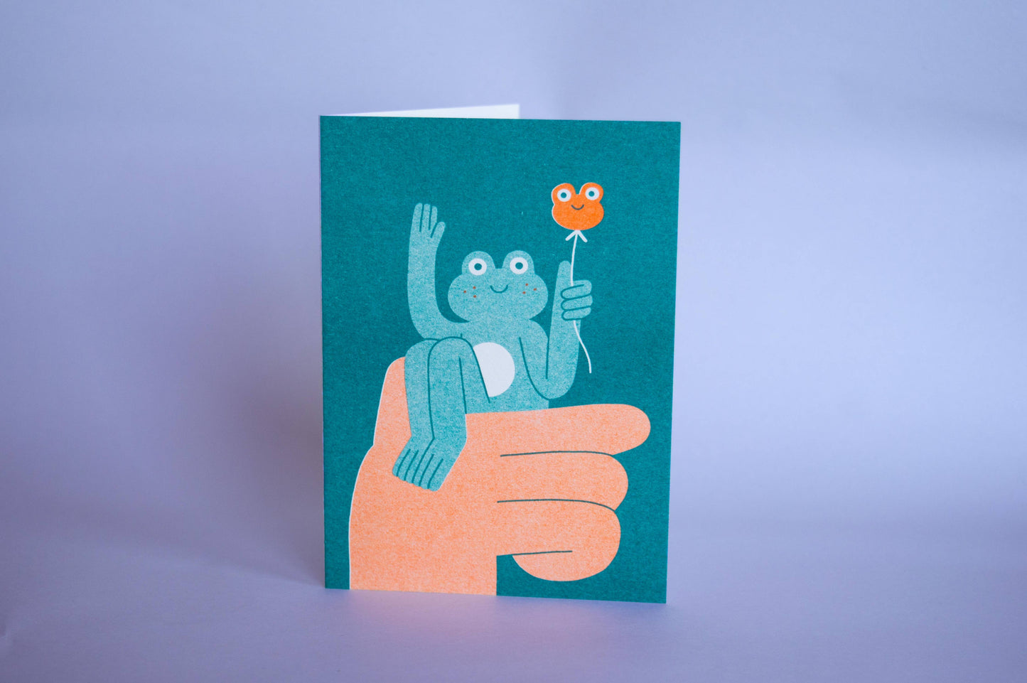 Hello Frog Card — illustration of a hand holding up a smiling frog with a frog-shaped balloon, shown on teal card stock