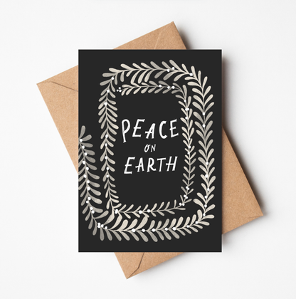 'Peace on Earth' Christmas card featuring a black background bordered by a cream botanical wreath, with 'PEACE ON EARTH' hand-lettered in the centre