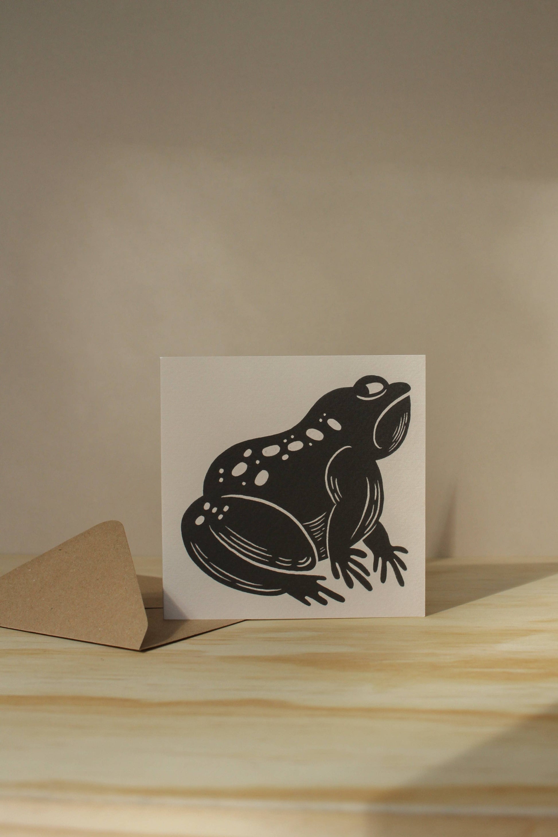 'The Grumpy Frog' card shown at an angle on its kraft envelope, part of Lauren Marina's square animal card series