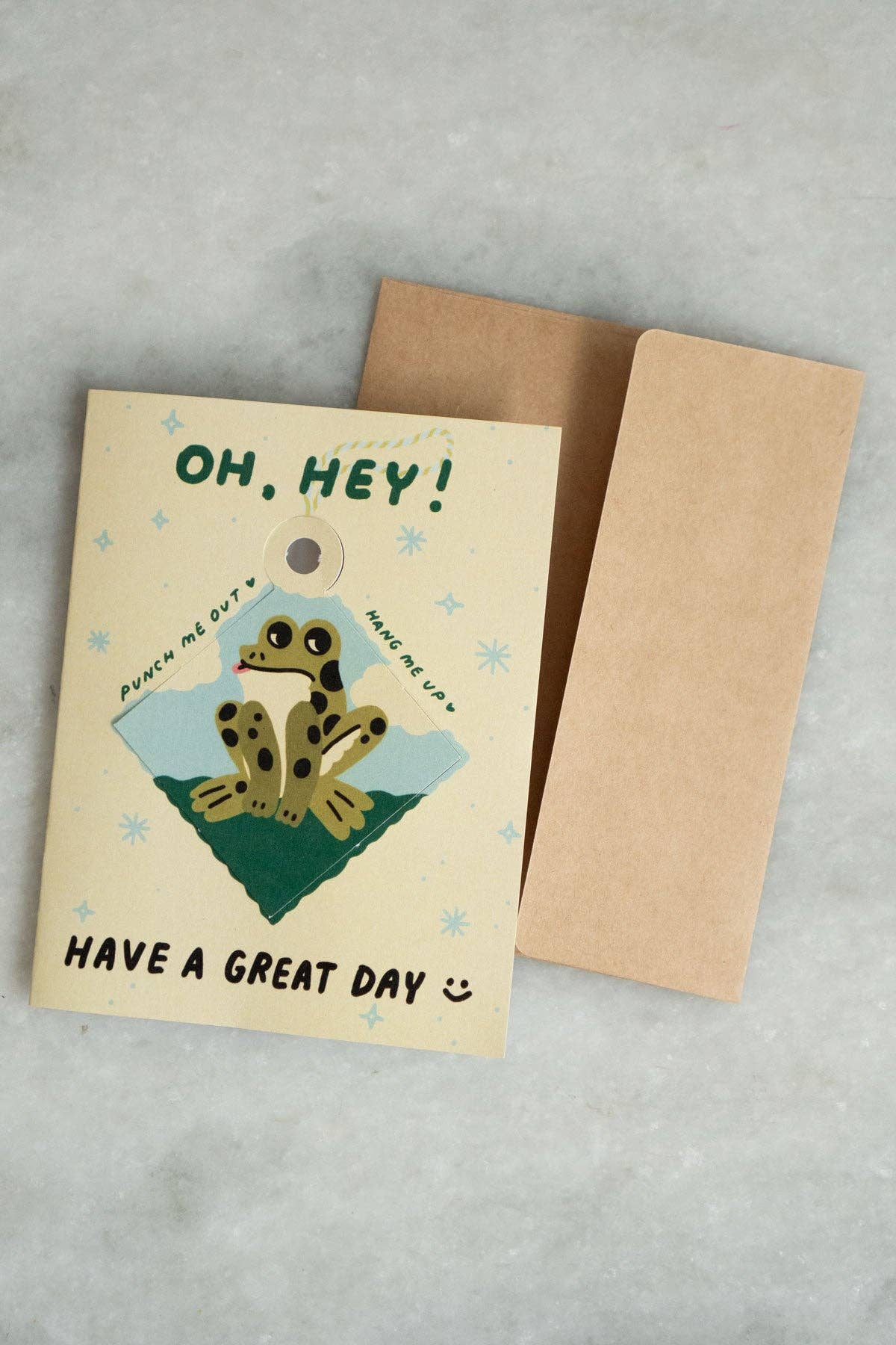 Cream 'Oh, Hey! Have a Great Day' greeting card with a punch-out diamond ornament featuring an illustrated spotted frog on a green hill, kraft envelope alongside