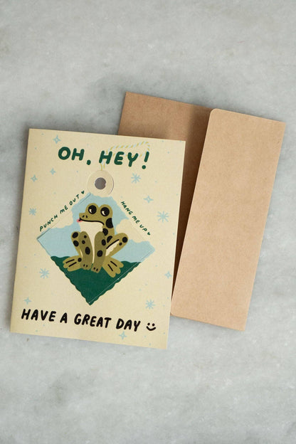 Cream 'Oh, Hey! Have a Great Day' greeting card with a punch-out diamond ornament featuring an illustrated spotted frog on a green hill, kraft envelope alongside