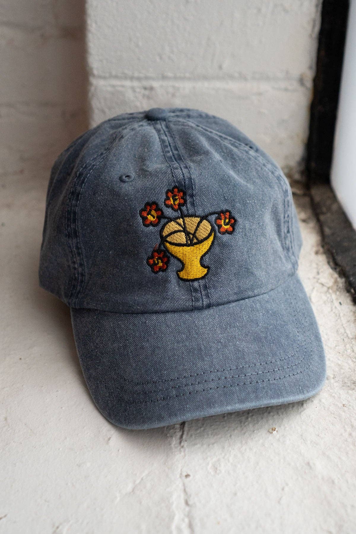 Vintage dark blue-grey dad hat with an embroidered flower vase design where the blooms spell out a hidden message, on a stone doorway ledge