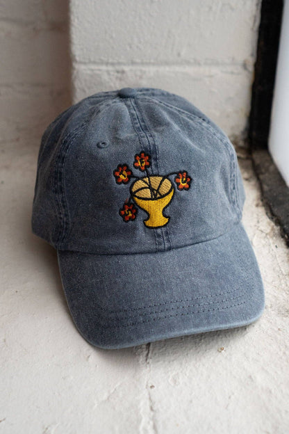 Vintage dark blue-grey dad hat with an embroidered flower vase design where the blooms spell out a hidden message, on a stone doorway ledge