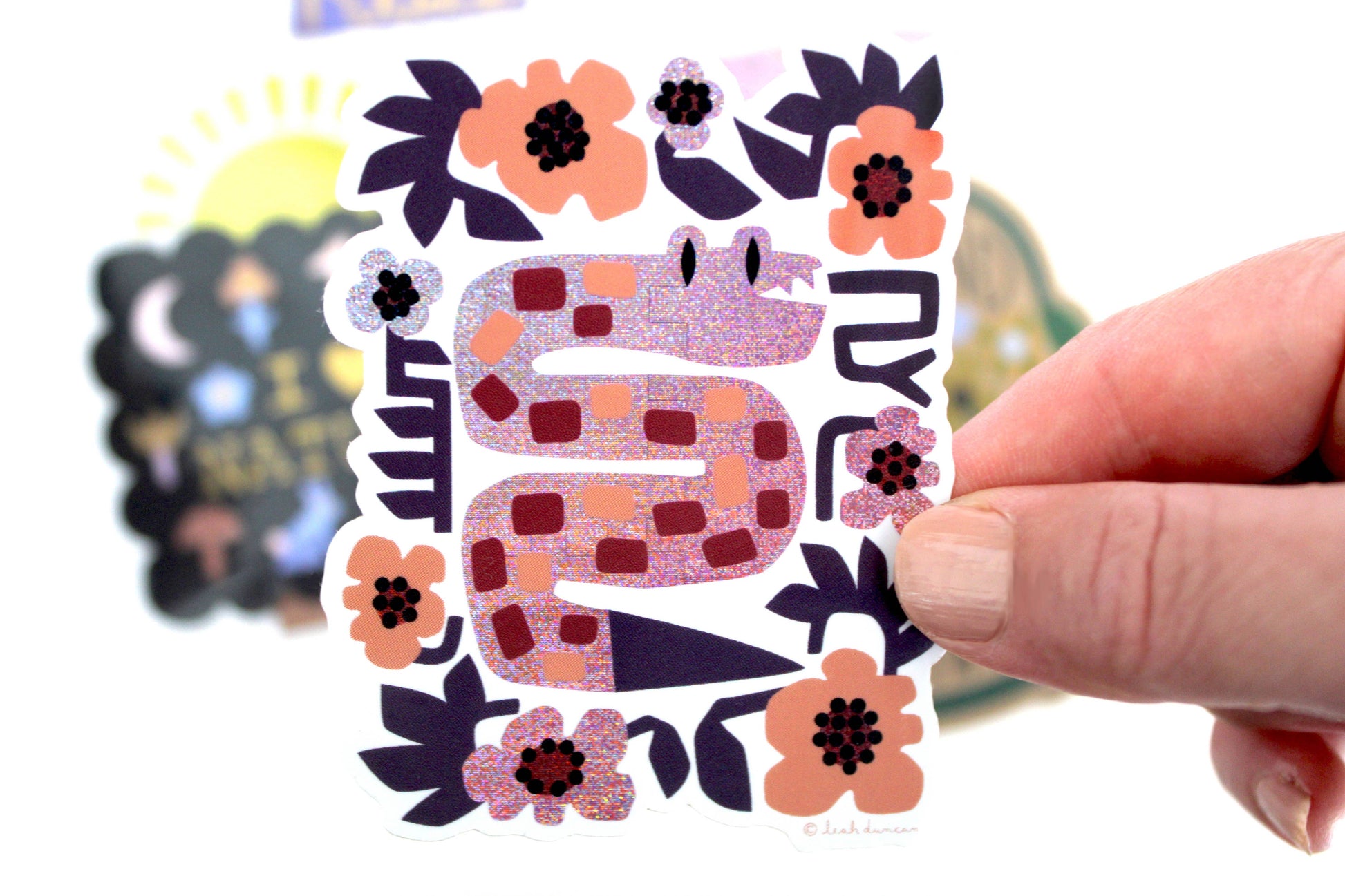 Glittery lavender snake vinyl sticker surrounded by purple and burgundy flowers, held in hand — PABOOM