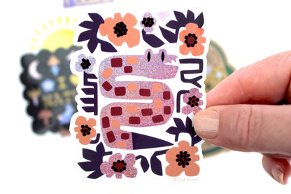 Glittery lavender snake vinyl sticker surrounded by purple and burgundy flowers, held in hand — PABOOM