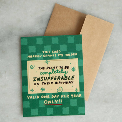 Green checkered greeting card reading 'This Card Hereby Grants Its Holder the Right to Be Completely Insufferable on Their Birthday, Valid One Day Per Year Only,' kraft envelope alongside