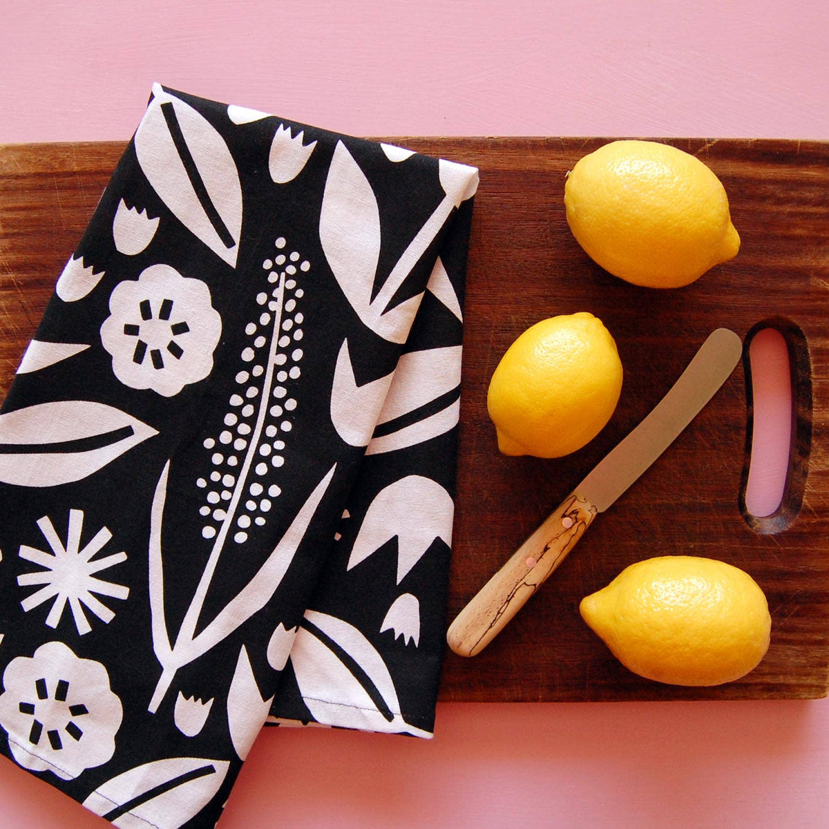 Black tea towel with white screen printed florals folded on a wooden cutting board beside lemons - PABOOM