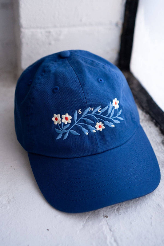 Royal blue dad hat with an embroidered floral garland design and 'SHC' lettering, propped against a white doorway