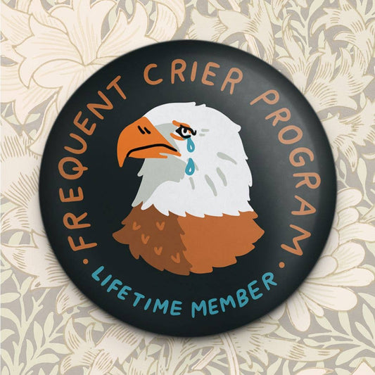 Round black fridge magnet with an illustrated crying bald eagle and 'Frequent Crier Program Lifetime Member' text, on a floral wallpaper background