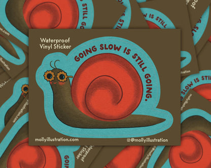 Matte vinyl sticker of a teal and orange cartoon snail with the text 'Going Slow Is Still Going' on a brown background