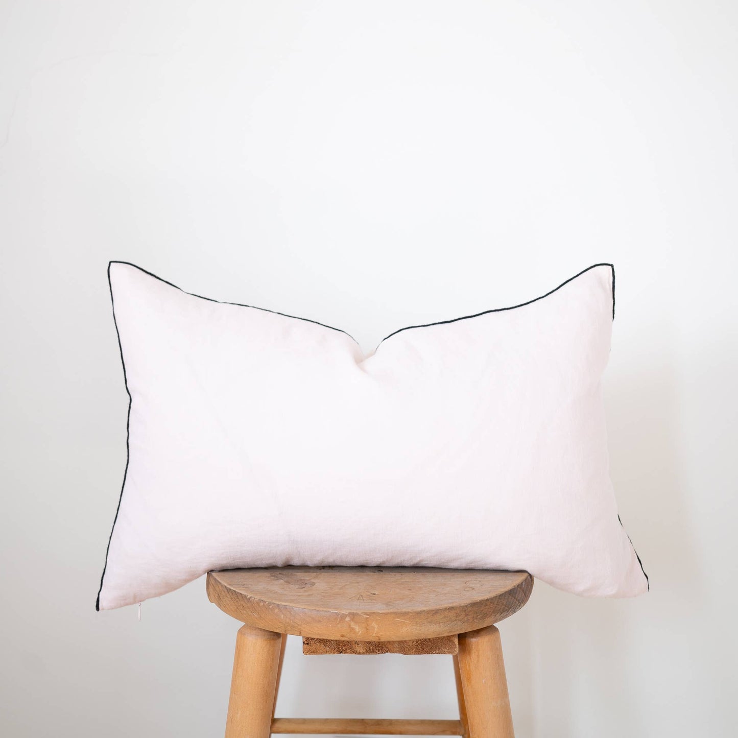 Blush pink 12x20 linen lumbar pillow cover with black contrast-edge trim displayed on a wooden stool against a white wall
