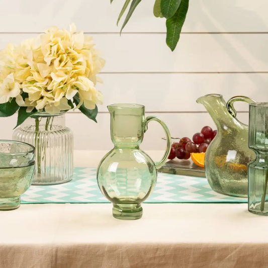 Recycled glass pitcher with a bulge middle and handle, green. On table with other glass products- PABOOM
