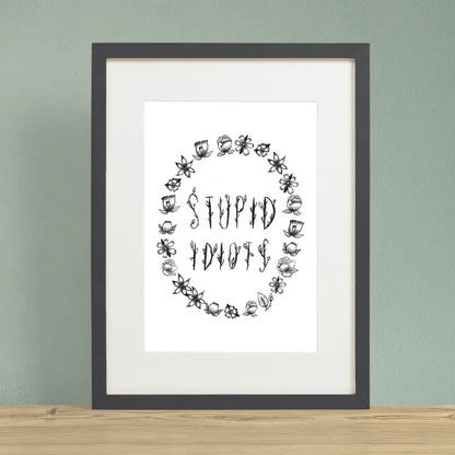 Floral black font saying STUPID IDIOTS surrounded by flower outlines on a white background print in black frame - PABOOM
