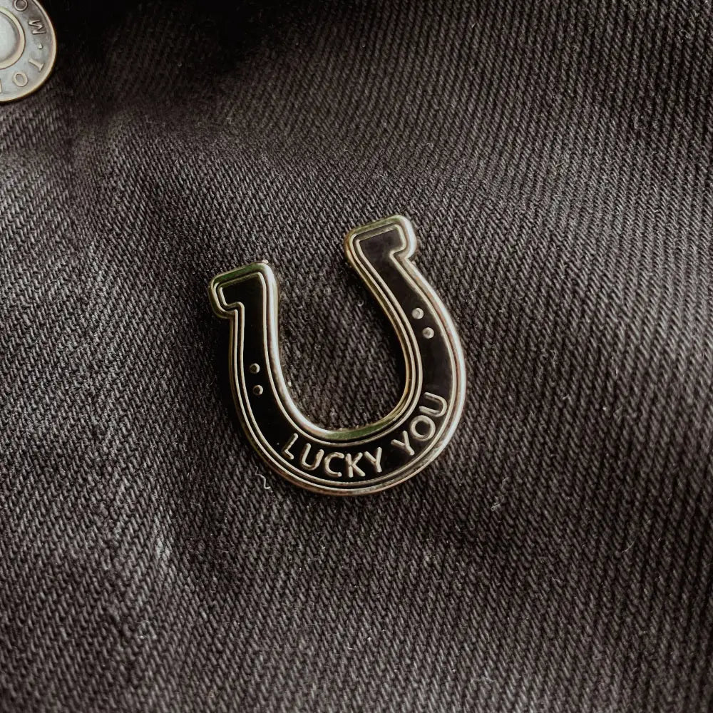 A black and silver lapel pin in the shape of a horseshoe with text "lucky you" on fabric