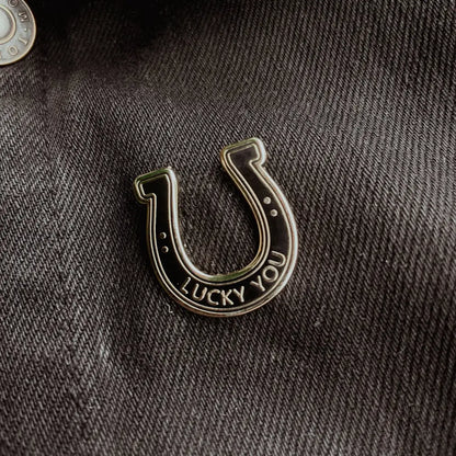 A black and silver lapel pin in the shape of a horseshoe with text "lucky you" on fabric