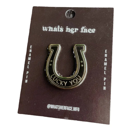 A black and silver enamel lapel pin in the shape of a horseshoe with text "lucky you" on black cardstock