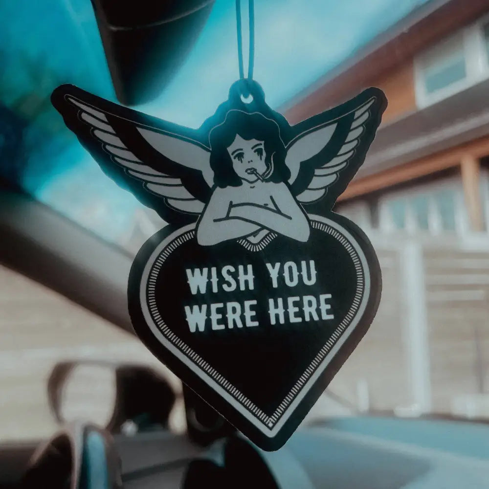 Airfreshener hanging in car of an angel smoking and leaning on heart saying Wish you were here